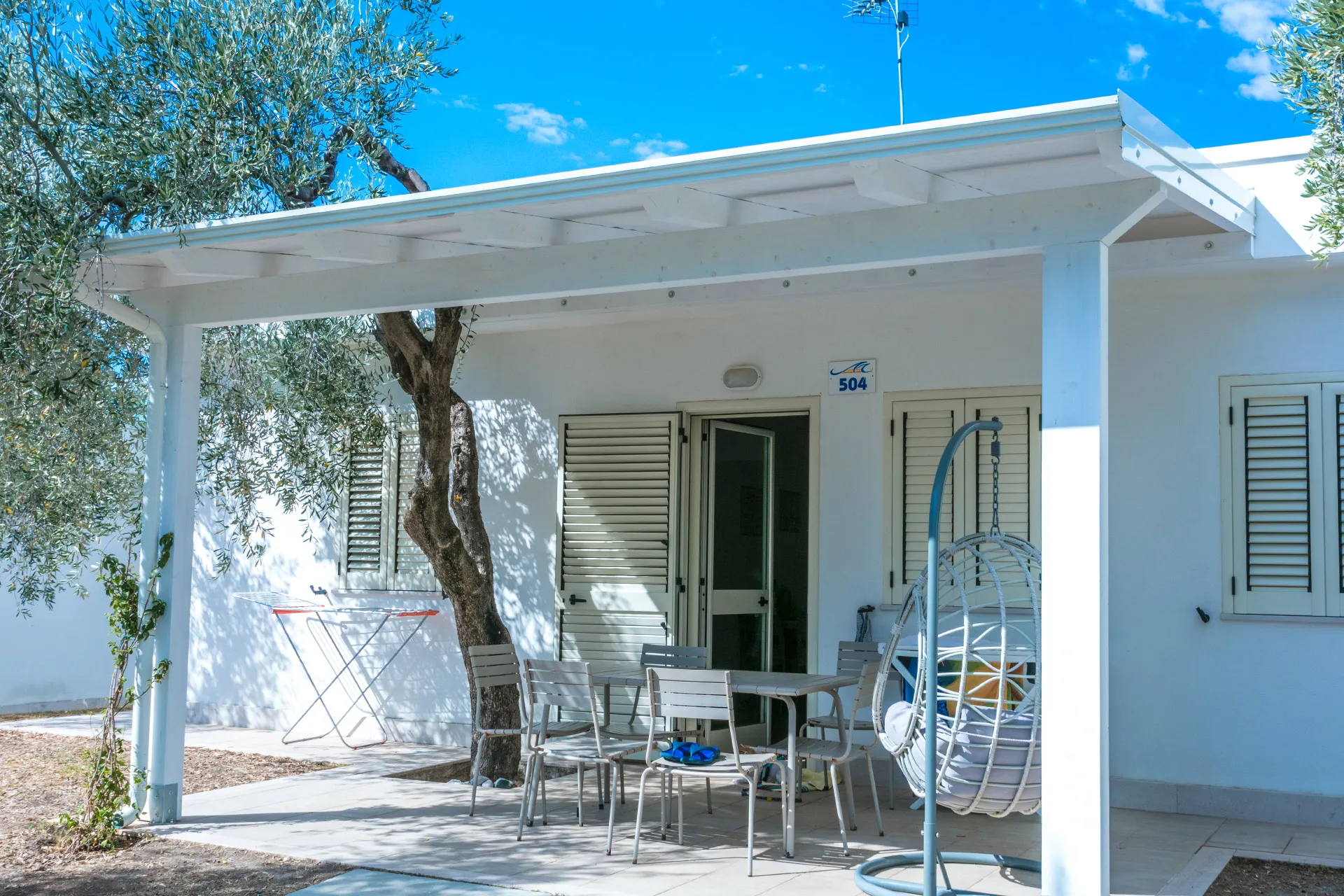 Veranda di bungalow con tavolo, sedie e poltrona sospesa all'ombra di un ulivo