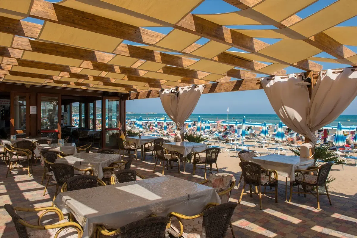 Terrazza ristorante con pergolato in legno e tessuto, tavoli apparecchiati e vista sulla spiaggia