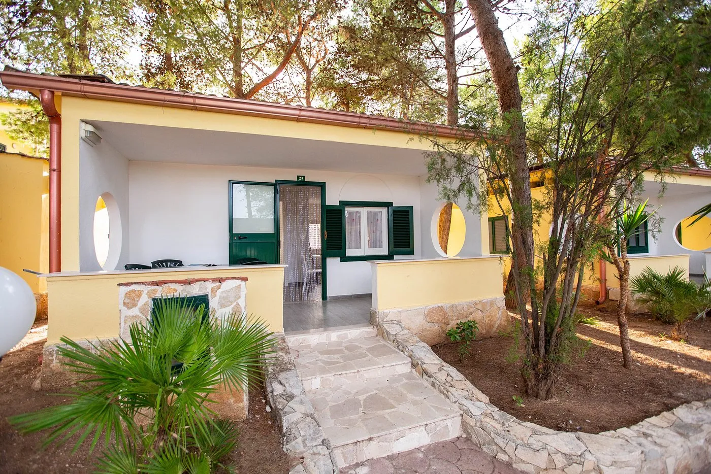 Esterno di bungalow giallo e bianco con portico, persiane verdi e vialetto in pietra tra i pini