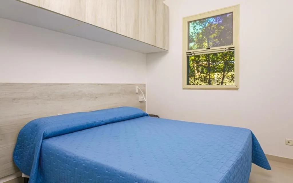 Camera da letto con copriletto azzurro, mobili in legno chiaro e finestra sul verde