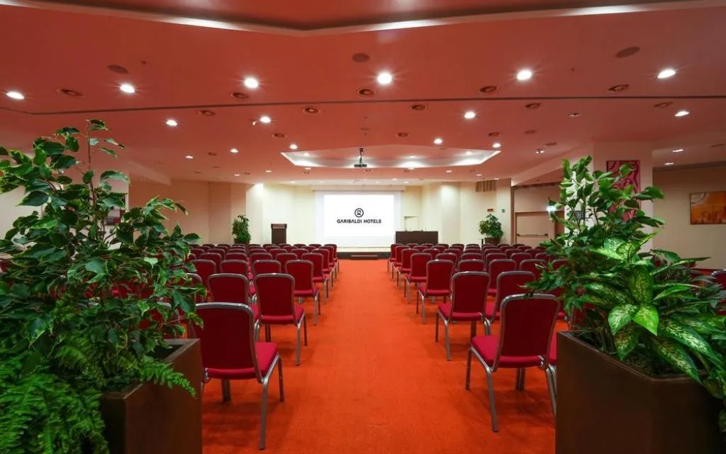 Ampia sala conferenze con sedie rosse, schermo per proiezioni e piante decorative