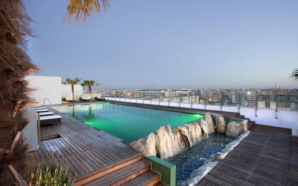 Piscina panoramica sul tetto con deck in legno, cascata di rocce e vista sulla città al crepuscolo