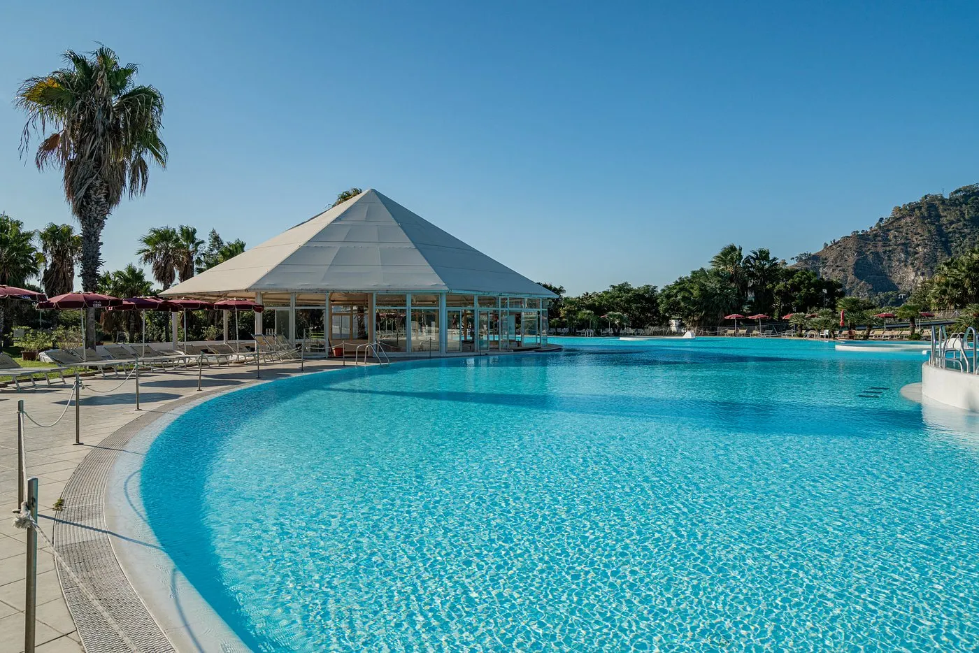 Piscina azzurra con gazebo a piramide e palme sotto cielo limpido