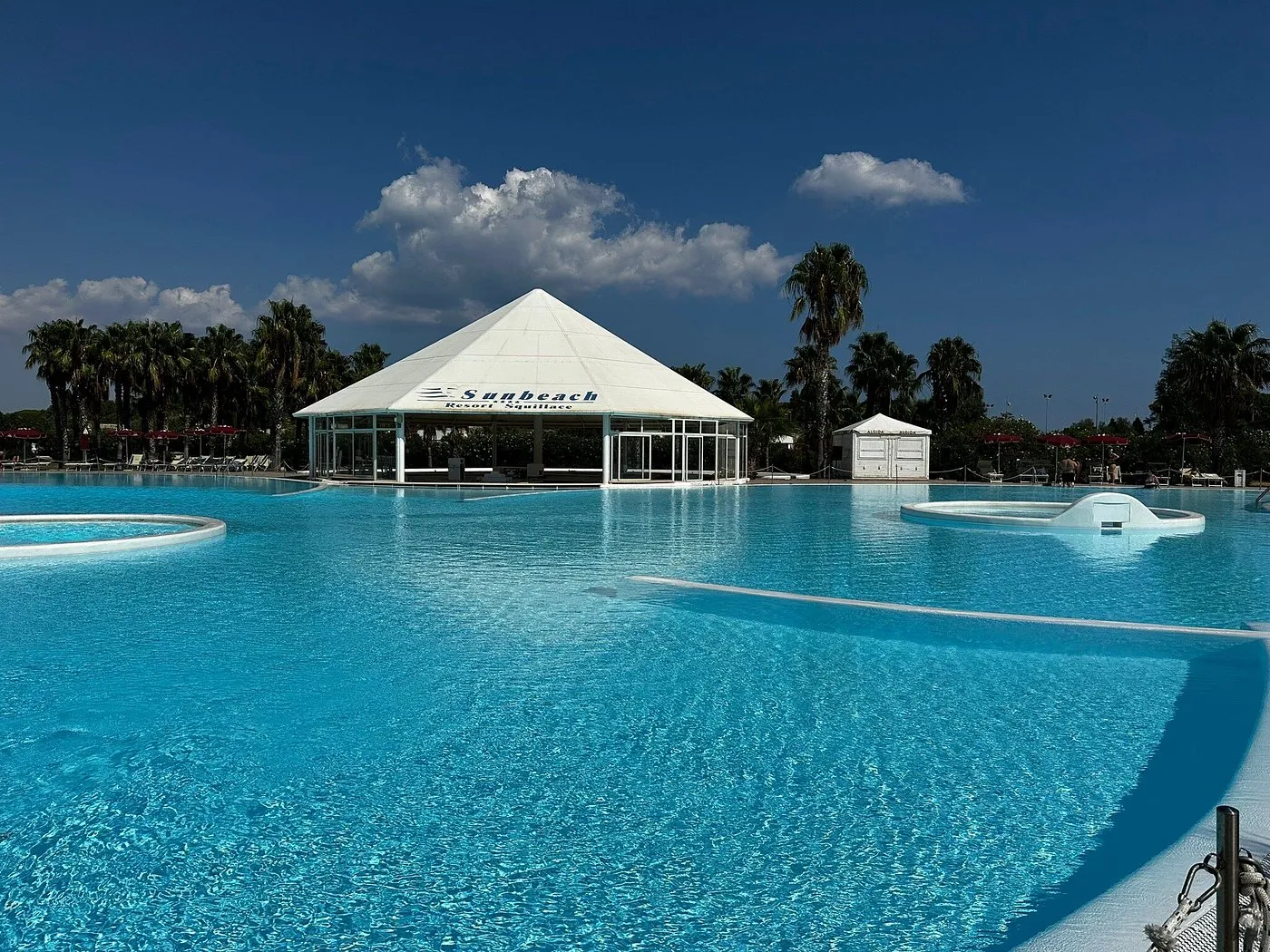 Piscina con gazebo bianco e palme sotto cielo azzurro con nuvole