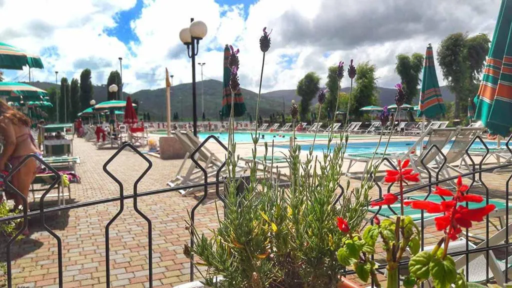 Area piscina vista attraverso una recinzione con piante e fiori, lettini e ombrelloni