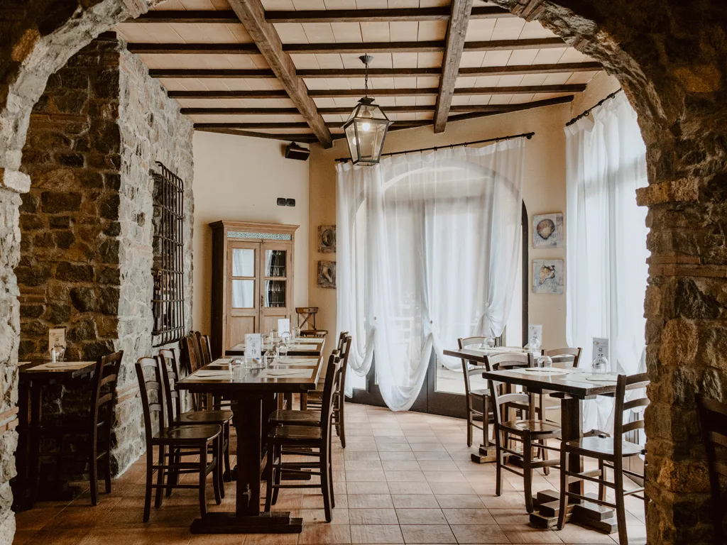 Sala ristorante rustica con muri in pietra, archi, travi a vista e tende bianche