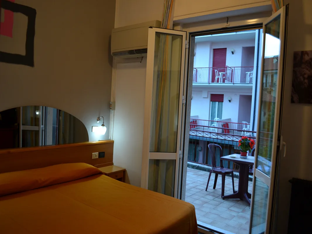 Camera con letto arancione e porta-finestra aperta sul balcone con tavolino e sedie