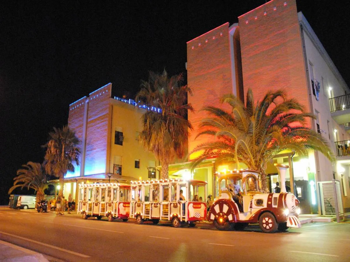 Vista notturna dell'hotel illuminato con trenino turistico e palme