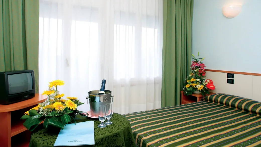 Camera d'albergo con letto matrimoniale, tende verdi, televisore e tavolino con fiori e spumante