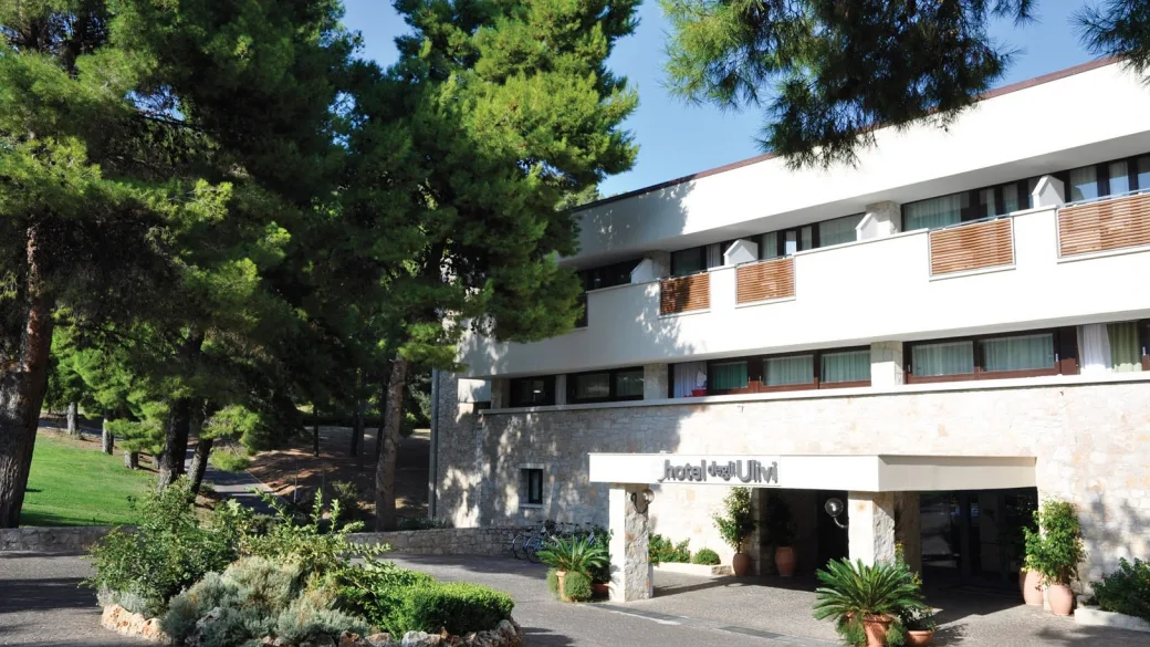 Ingresso dell'Hotel degli Ulivi con facciata bianca e in pietra circondata da pini e piante in vaso