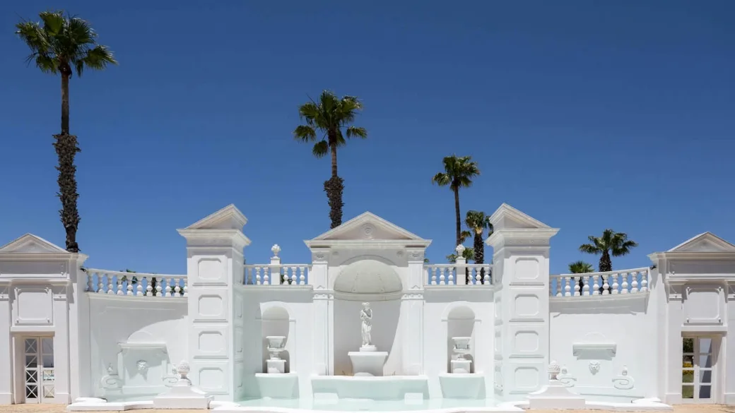 Fontana bianca monumentale con statue e palme sotto cielo azzurro