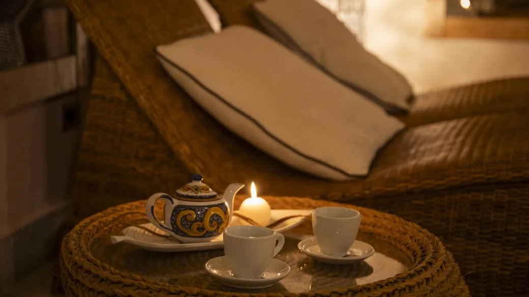 Dettaglio relax con teiera decorata, tazze e candela su tavolino in vimini