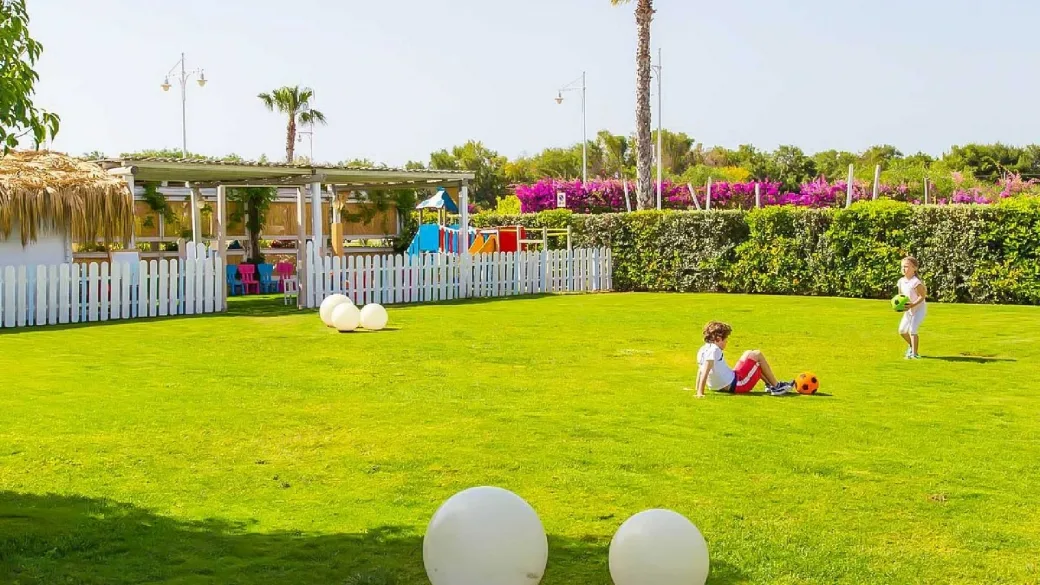 Prato verde con area giochi per bambini, recinzione bianca e bambini che giocano a pallone