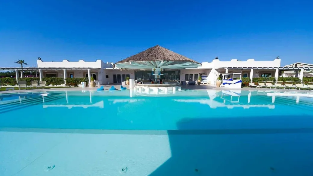 Ampia piscina con acqua turchese e edificio principale bianco con bar coperto da tetto in paglia