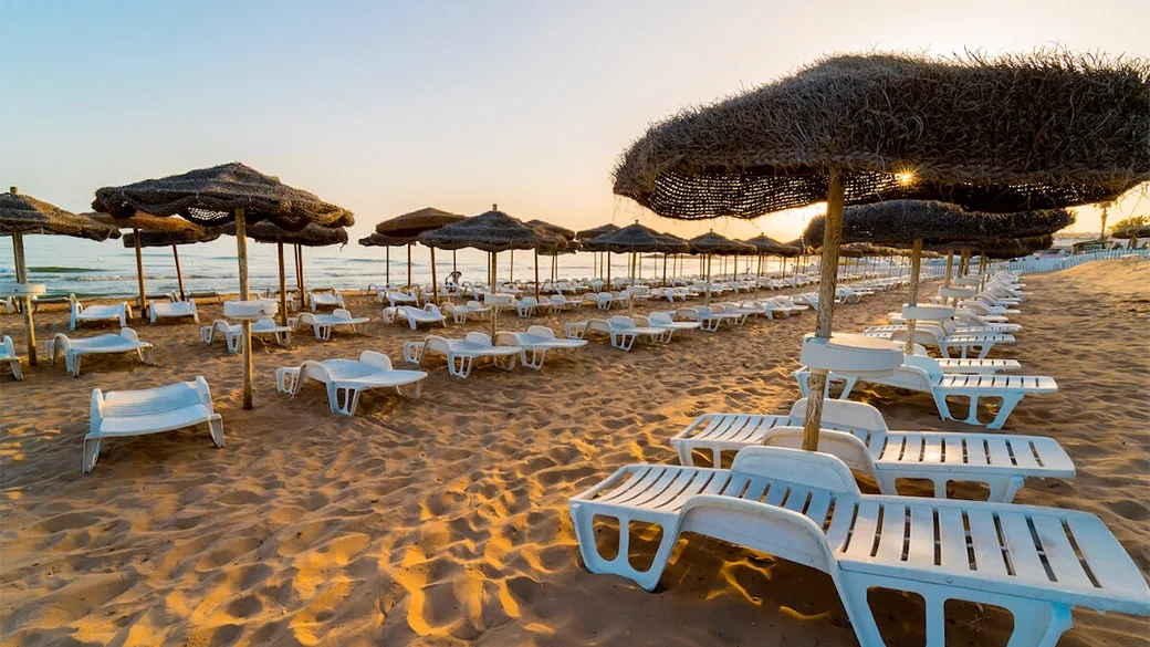 Spiaggia al tramonto con file di lettini bianchi e ombrelloni in paglia
