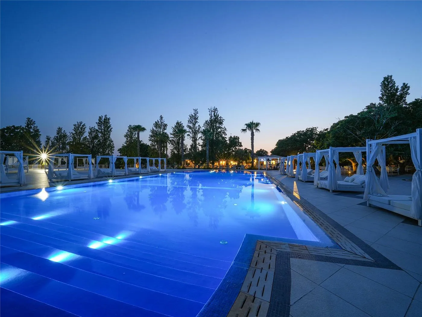 Piscina illuminata al tramonto con gazebo bianchi e palme