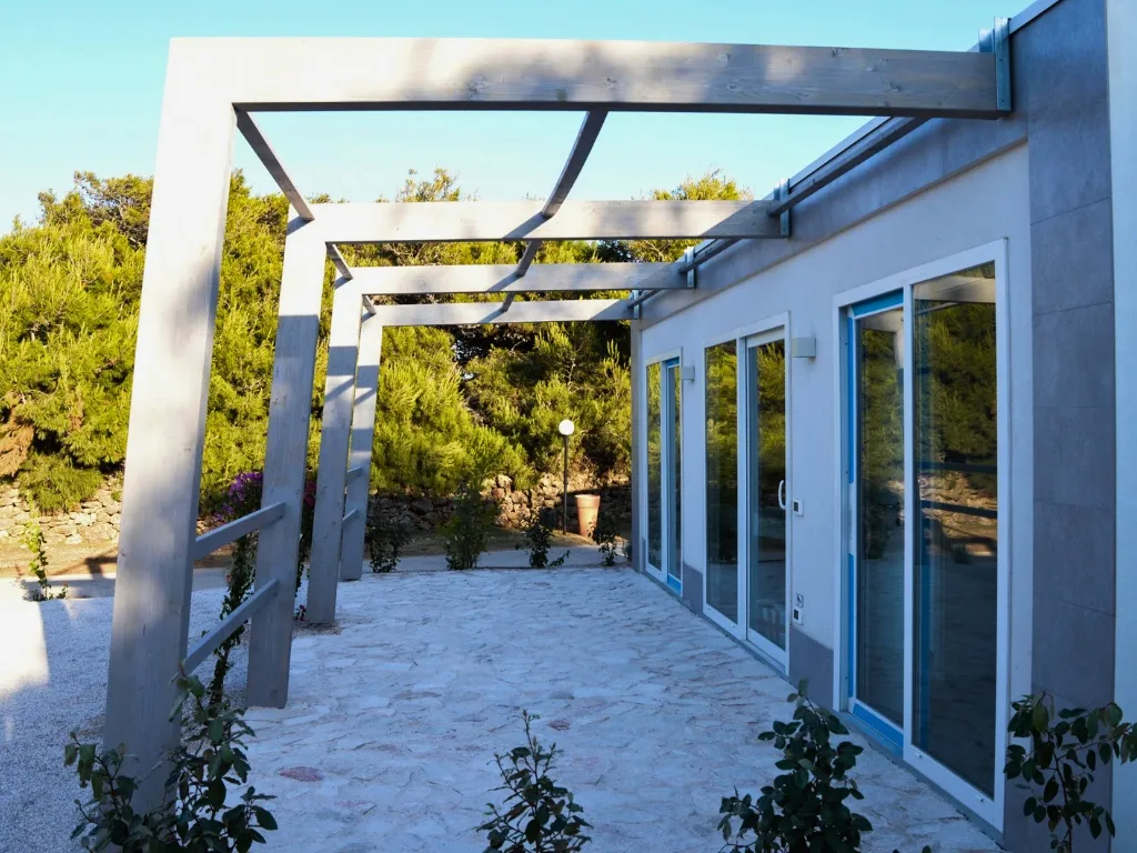Veranda esterna con pergolato in legno grigio e ampie vetrate su giardino roccioso