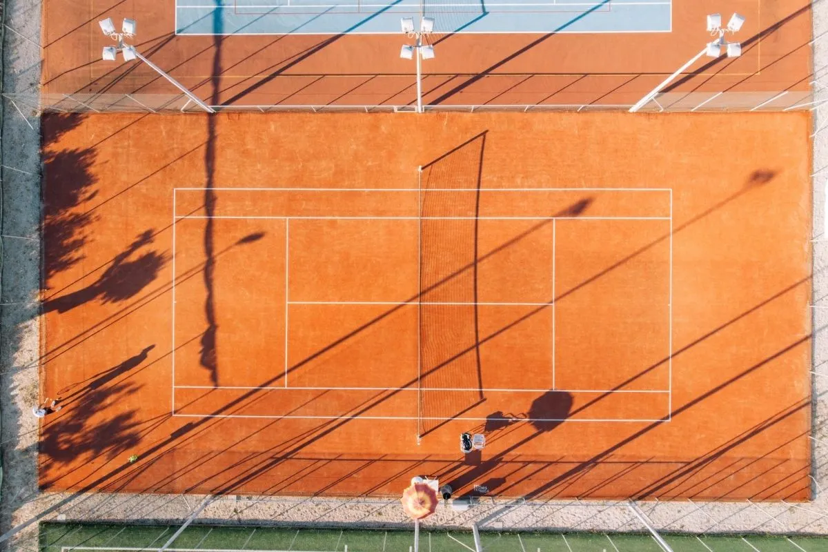 Campo da tennis in terra rossa visto dall'alto con ombre di palme