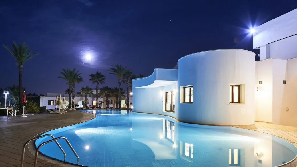 Piscina illuminata di notte accanto a edificio bianco con luna e palme