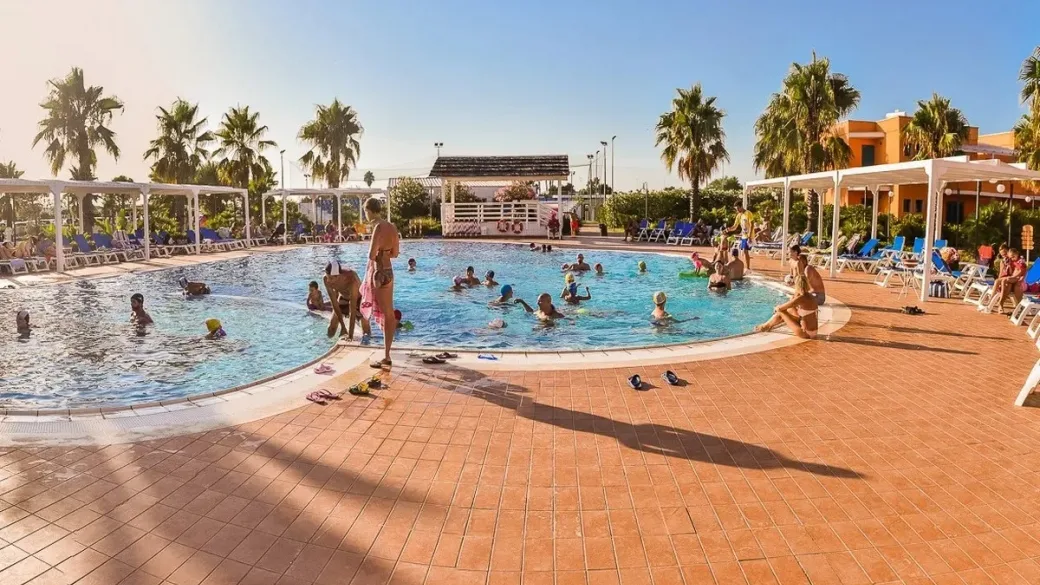 Piscina con bagnanti, lettini e palme in giornata di sole