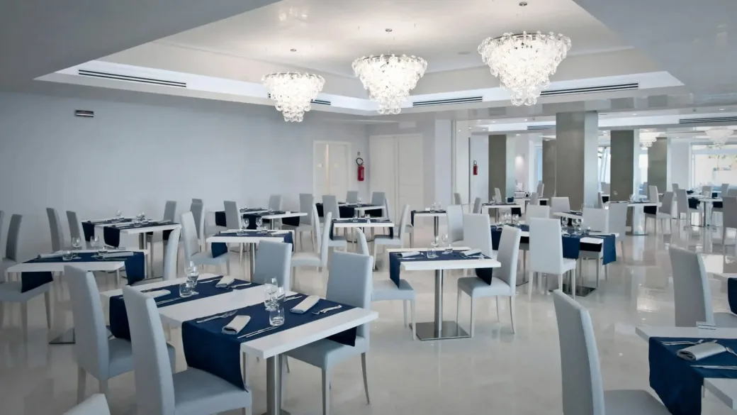 Ampia sala da pranzo luminosa con tovaglie blu, sedie bianche e lampadari di cristallo