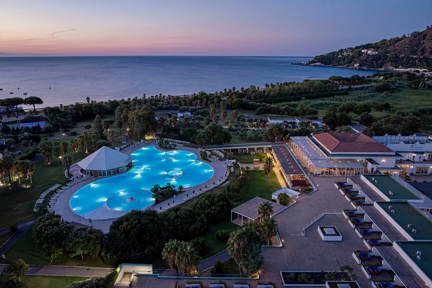 Vista aerea del resort al tramonto con piscina illuminata e mare sullo sfondo