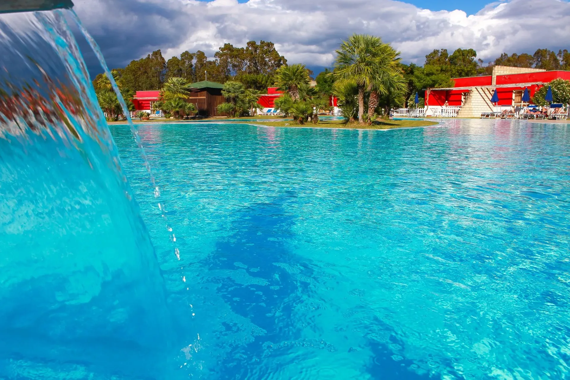 Piscina con cascata d'acqua e isolette di palme, edifici rossi del resort sullo sfondo