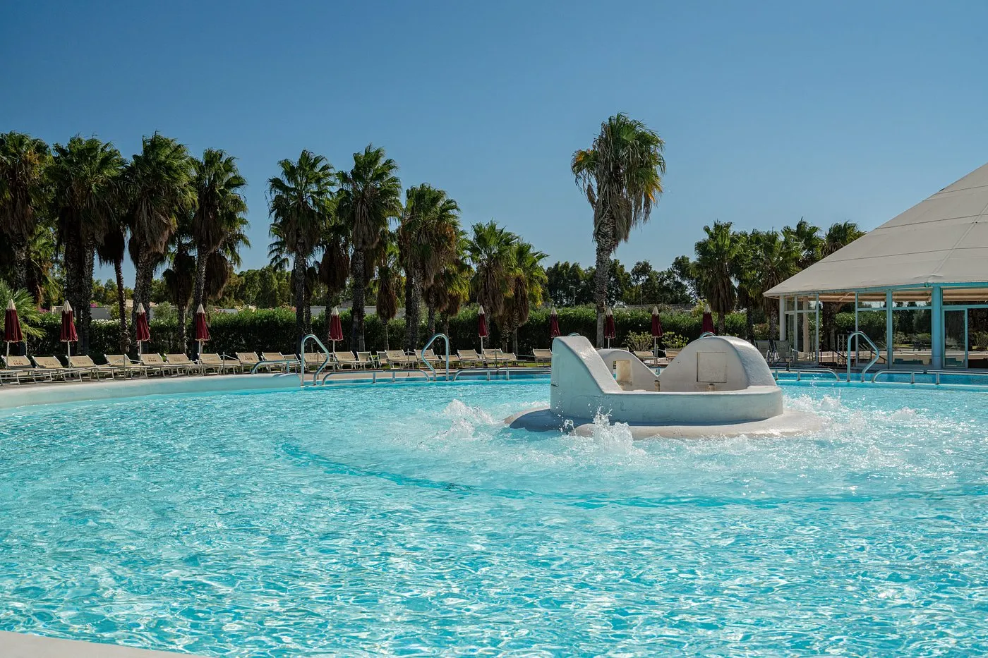 Dettaglio della piscina con giochi d'acqua e isola centrale, palme e lettini intorno