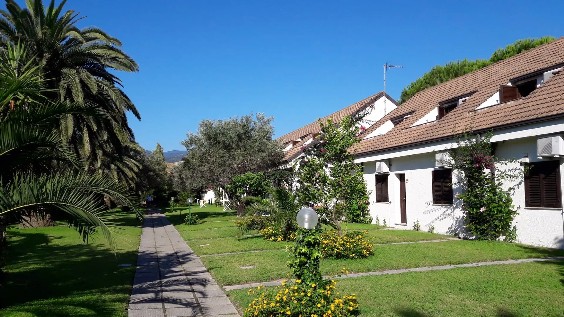 Vialetto tra gli alloggi bianchi del villaggio con giardini fioriti e palme