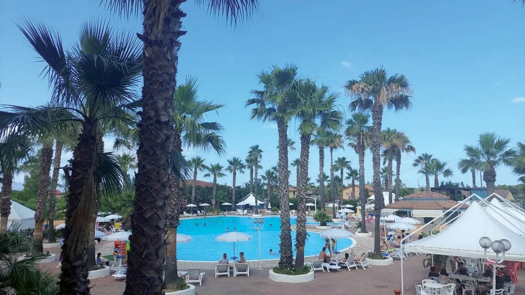 Piscina del resort vista tra le palme con ombrelloni, lettini e ospiti in una giornata di sole