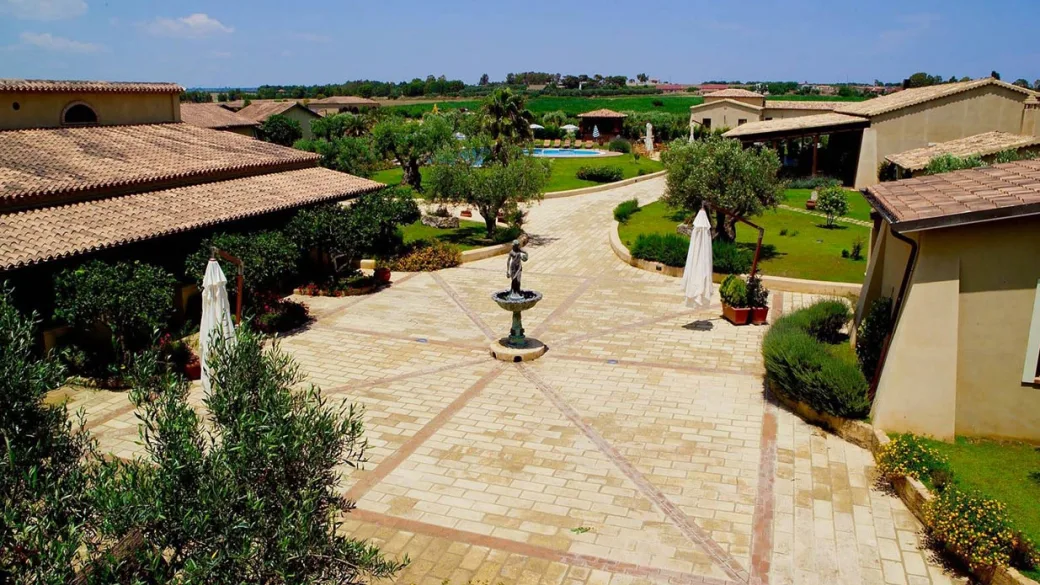 Cortile pavimentato con fontana centrale, ulivi e edifici in stile rustico