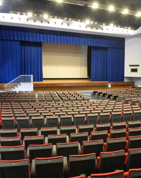 Interno di un auditorium con palco, quinte blu e file di poltrone rosse e nere vuote.