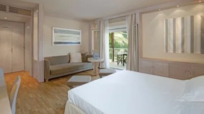 Camera d'hotel luminosa con letto matrimoniale, divano, tavolino, terrazza con sedie e vista su palme verdi.