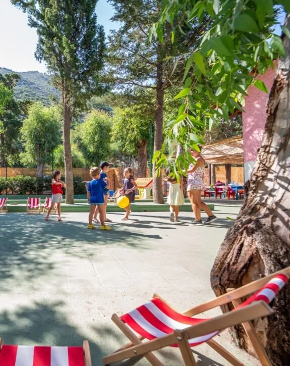 Bambini che giocano all'aperto in un parco ombreggiato con alberi, sedie a sdraio rosse e bianche e montagne sullo sfondo.