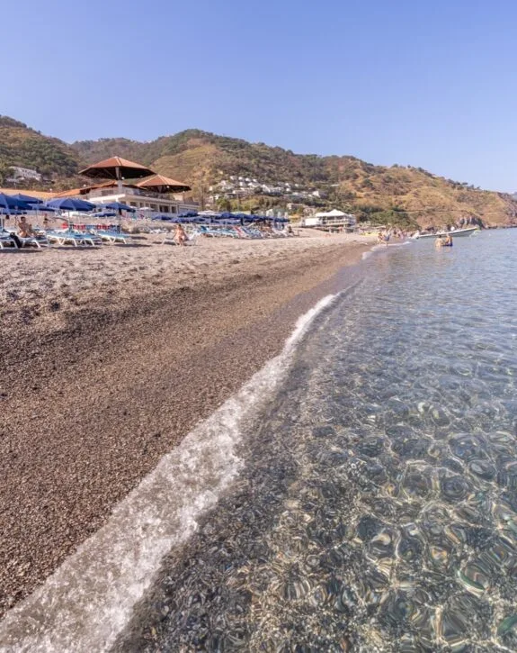 Spiaggia con ghiaia, ombrelloni blu e sdraio, mare cristallino e colline verdeggianti sullo sfondo.