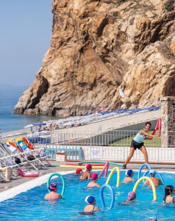 Lezione di acquagym con istruttrice e partecipanti con cuffie colorate in piscina sullo sfondo di una scogliera.