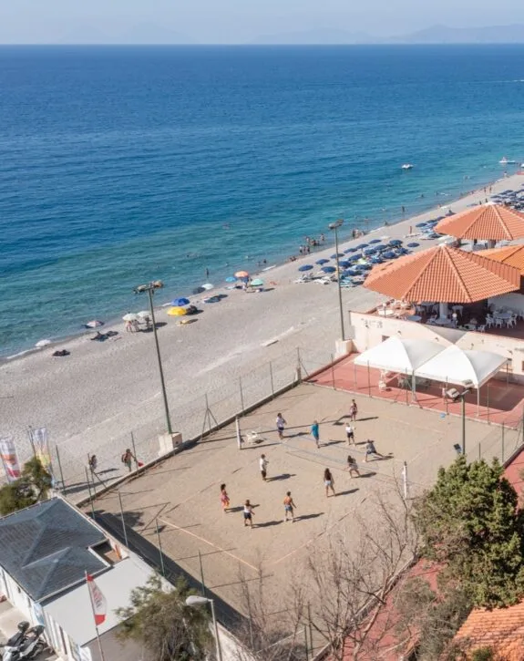 Spiaggia con ombrelloni, persone in acqua e campo da beach volley con giocatori in azione.