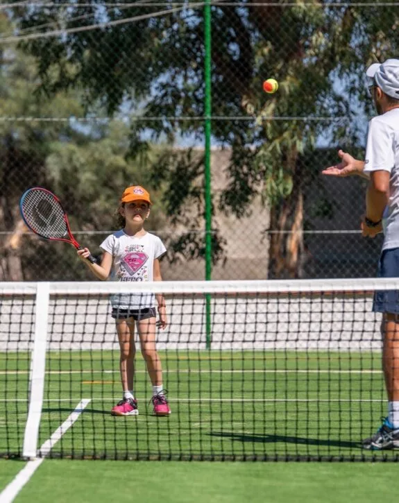 Bambina con cappellino arancione pronta a colpire la palla da tennis durante una lezione all'aperto su campo verde.