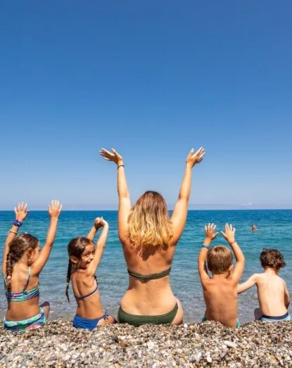 Donna e quattro bambini seduti su una spiaggia di ciottoli con le mani alzate verso il mare calmo e il cielo sereno.