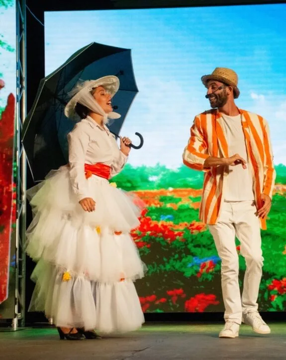 Due attori in costume colorato su un palco con sfondo floreale, lei con ombrello e lui con cappello di paglia.