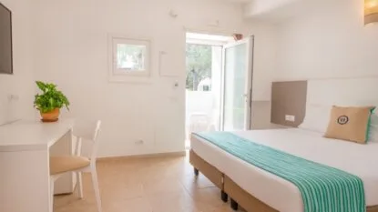 Camera d'albergo luminosa con letto matrimoniale, scrivania bianca, pianta, porta finestra aperta verso terrazza esterna.