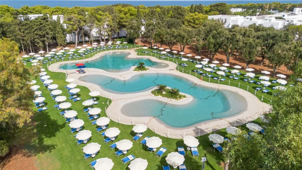Piscina a forma libera con ombrelloni bianchi e lettini blu su un prato verde, circondata da alberi e edifici bianchi.
