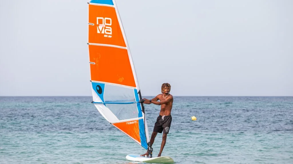 Uomo su tavola da windsurf con vela arancione e blu sul mare calmo e cielo limpido.