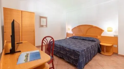 Camera da letto con letto matrimoniale, testiera in vimini, scrivania con TV e sedia rossa, armadio e pareti bianche.