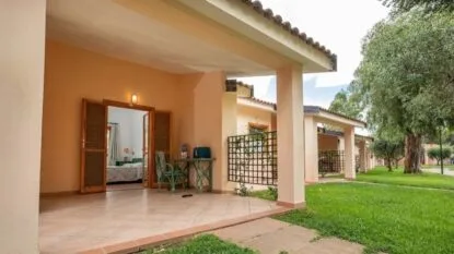 Veranda di una casa con tavolo e sedia verde, porte aperte su camera da letto, giardino verde e alberi.