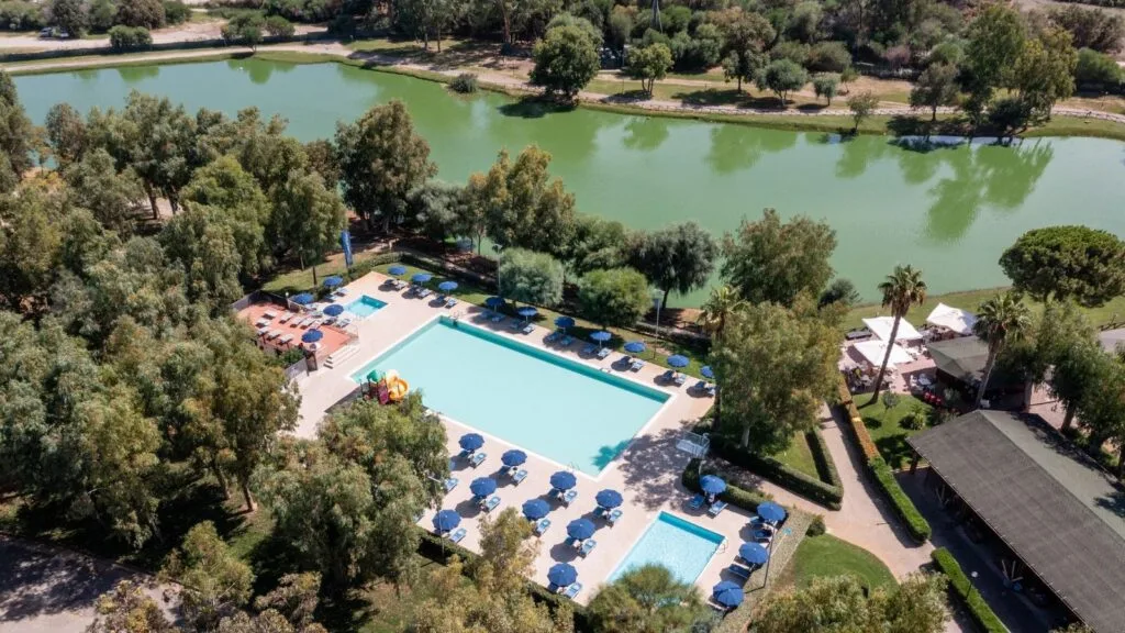 Piscina all'aperto con ombrelloni blu, circondata da alberi e un laghetto verde sullo sfondo.
