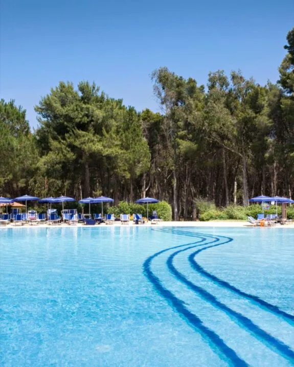 Piscina all'aperto con ombrelloni blu e lettini, circondata da alberi verdi e cielo sereno.