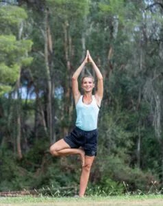 Donna sorridente in tenuta sportiva, pratica yoga all'aperto con posa dell'albero in un ambiente naturale verde.