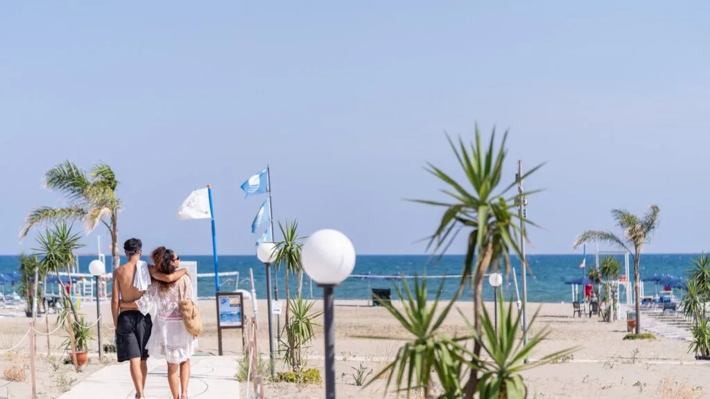 Coppia che cammina su passerella verso la spiaggia, mare e cielo blu sullo sfondo con palme e bandiere.