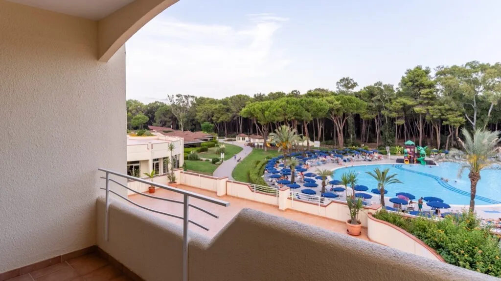 Vista da un balcone su piscina all'aperto con ombrelloni blu e area giochi, circondata da alberi verdi.